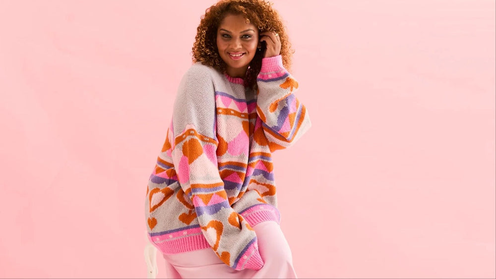 Woman wears Poppy Heart Intarsia Balloon Sleeve Oversized Jumper, against pink background