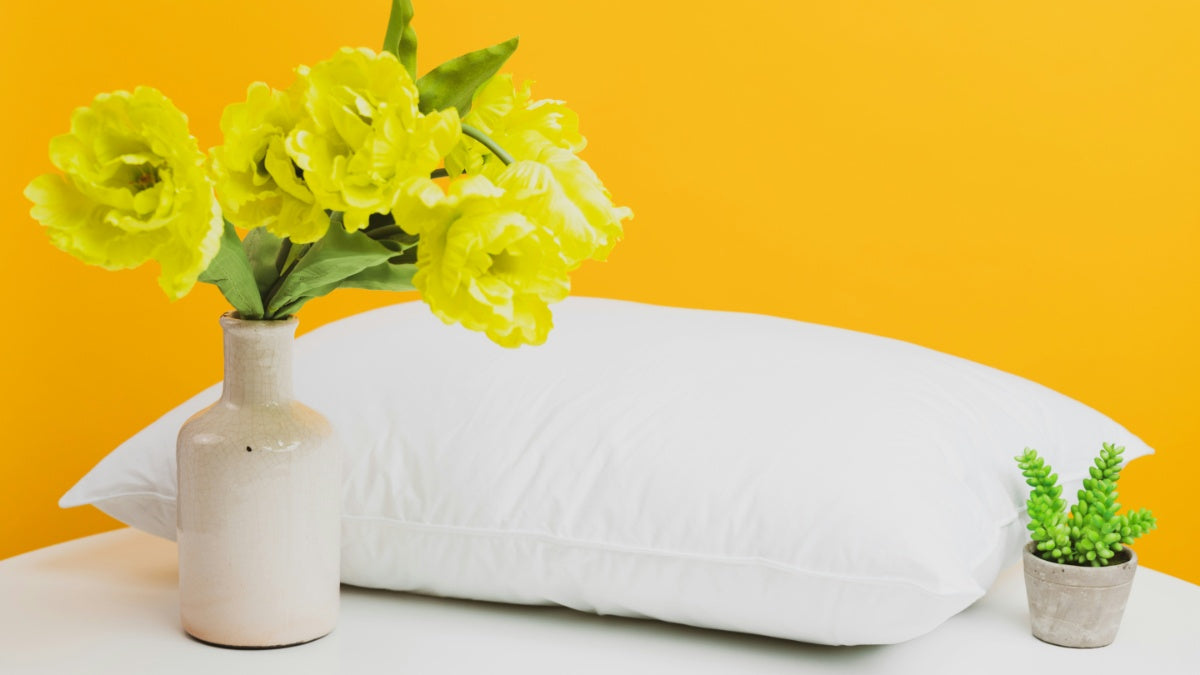 A white pillow against a yellow background next to a vase of flowers and  plant pot